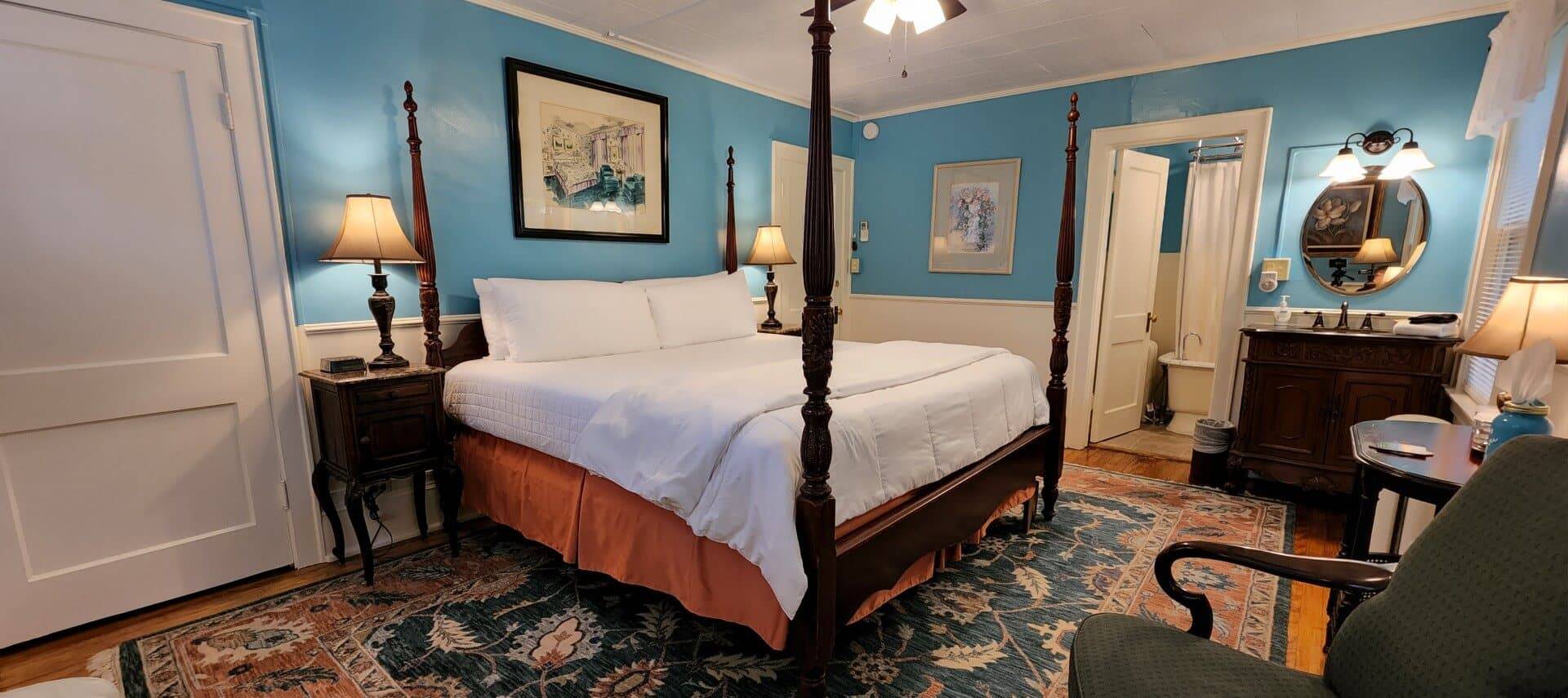 Bedroom with four poster bed, blue walls, antique furniture and doorway into a bathroom showing a clawfoot tub