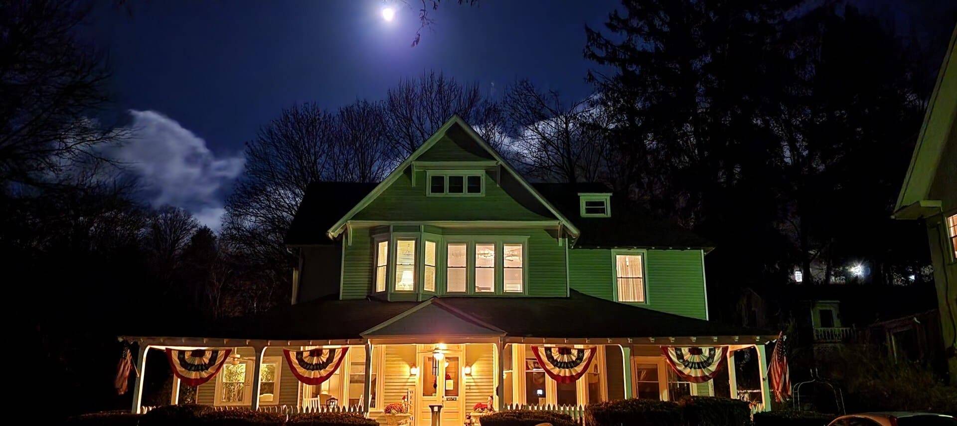Front of a home with large wrap around porch lit up at night with bright moon overhead