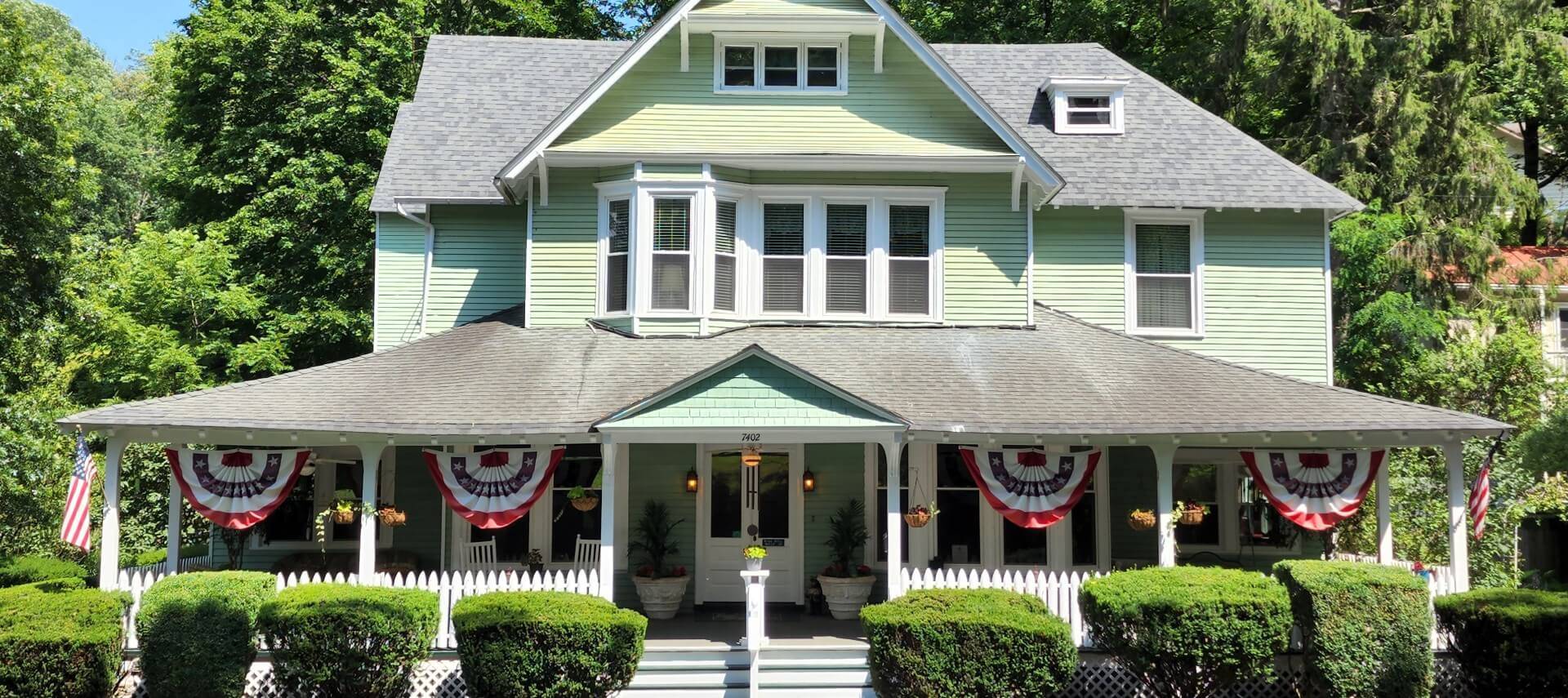 Large home with light green siding and wrap around porch with American bunting flags