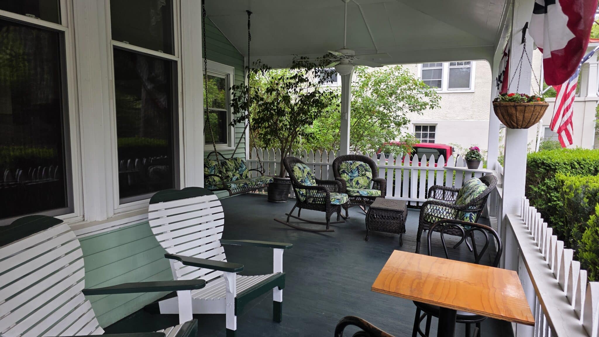 Cozy covered front porch with green floor, white Adirondack chairs, porch swing, wicker seating, and a wooden table.