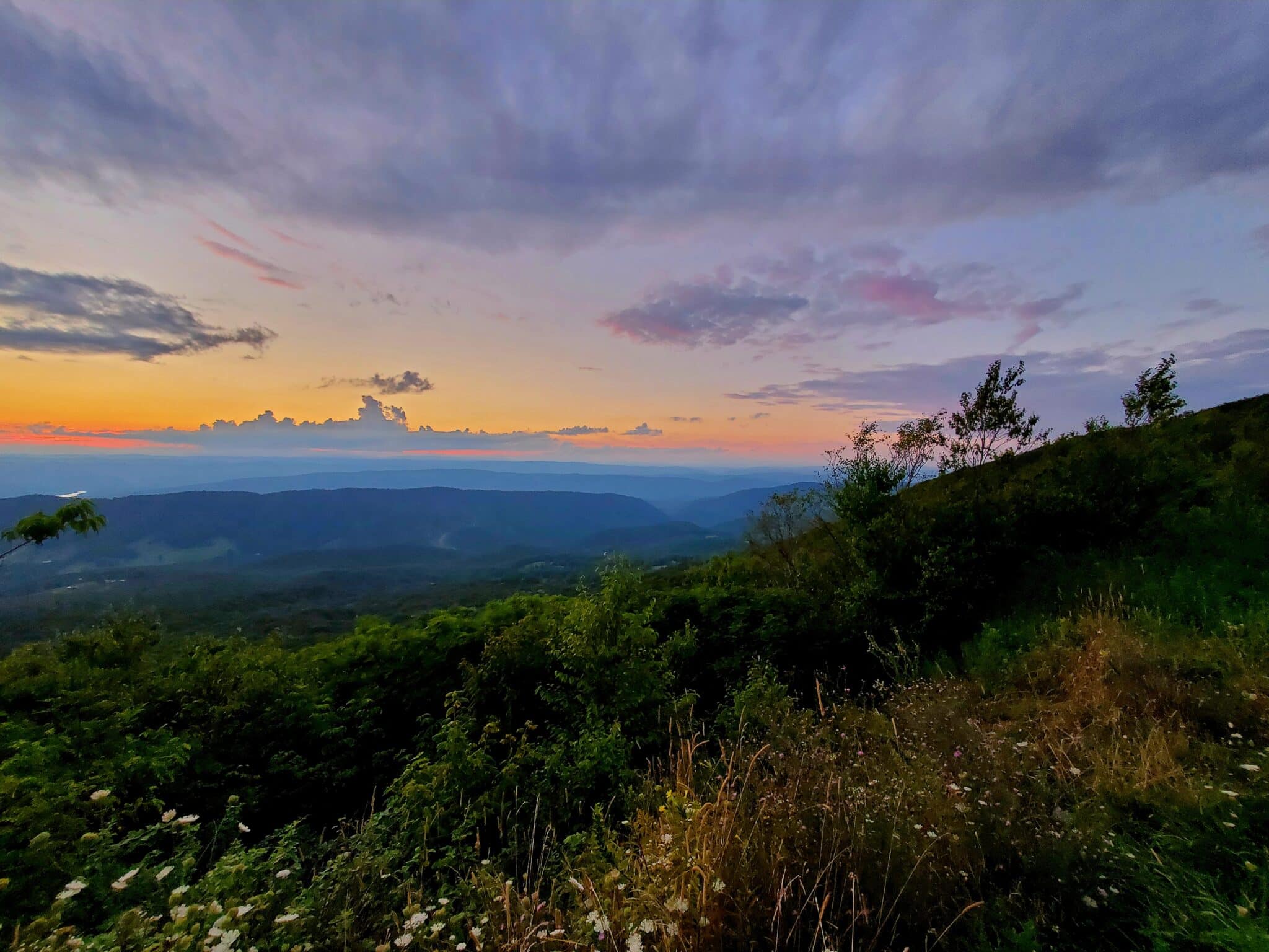 Sunset over rolling hills with an orange sky, purple clouds, and green foreground vegetation.