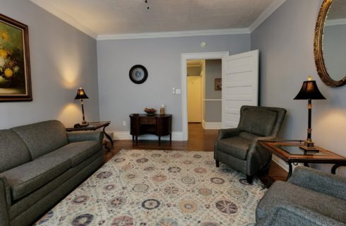 Sitting room with grey couch, two chairs tables with lamps and large round mirror on wall