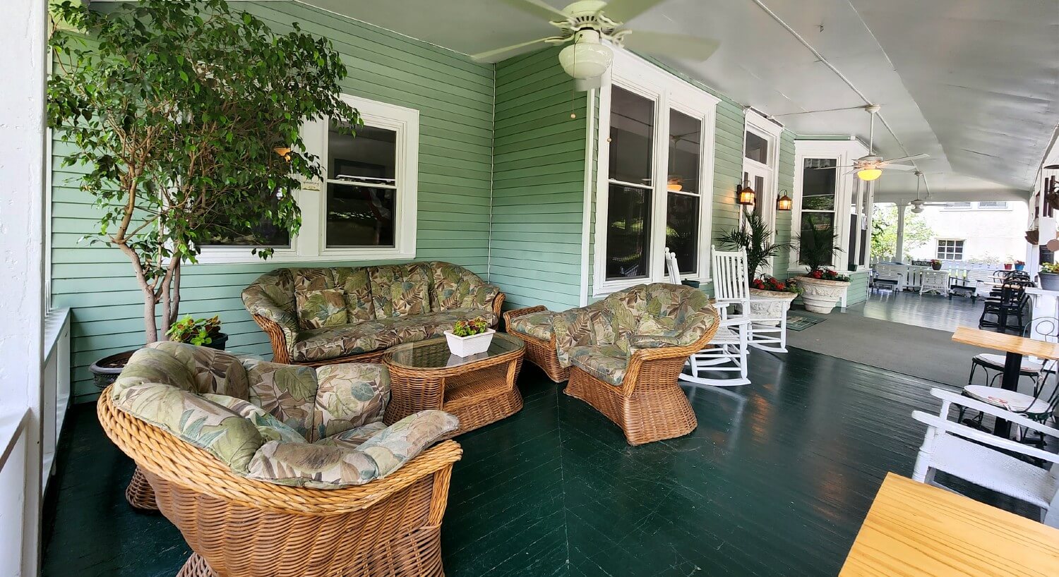 Expansive outdoor wrap around porch with wicker seating, plants and trees and white rocking chairs