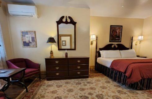Spacious guest room with bed, dark wood dresser with mirror and club chairs with a table and lamp