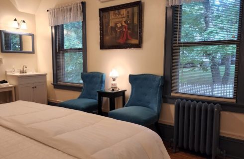 Guest room with white king bed, two blue sitting chairs by large windows and sink with mirror