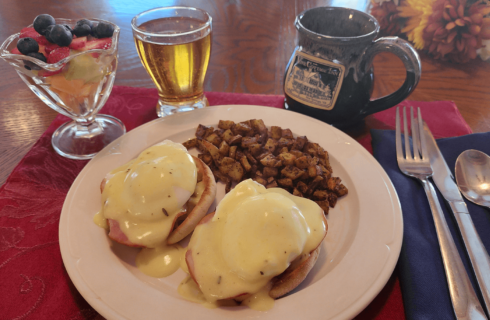 Breakfast plate of english muffin sandwiches with hollandaise sauce, potatoes and fruit, juice and coffee mug