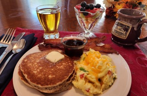 Breakfast plate of pancakes, eggs and bacon with juice, fruit and coffee mug