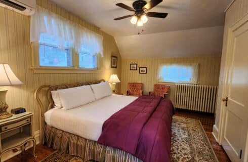 Guest room with striped wallpaper, king bed, nightstands with lamps, and striped sitting chairs. Large area rug over hardwood floors. Windows above bed and behind sitting chairs.
