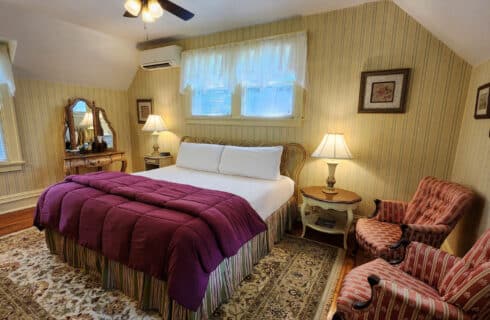 Elegant bedroom in soft cream colors with antique dresser, two nightstands with lamps, 2 striped armchairs. Windows on each wall, large area rug over hardwood floor.