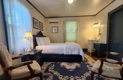 Guest room with bed with white linens, decorative blue rug and two antique sitting chairs