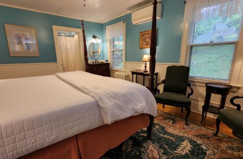 Guest room with four poster bed, sink vanity with round mirror and sitting chairs below a window