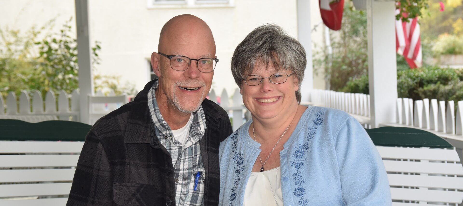 Woman in glasses and blue denim shirt sitting beside a man in plaid shirts on a white bench outside