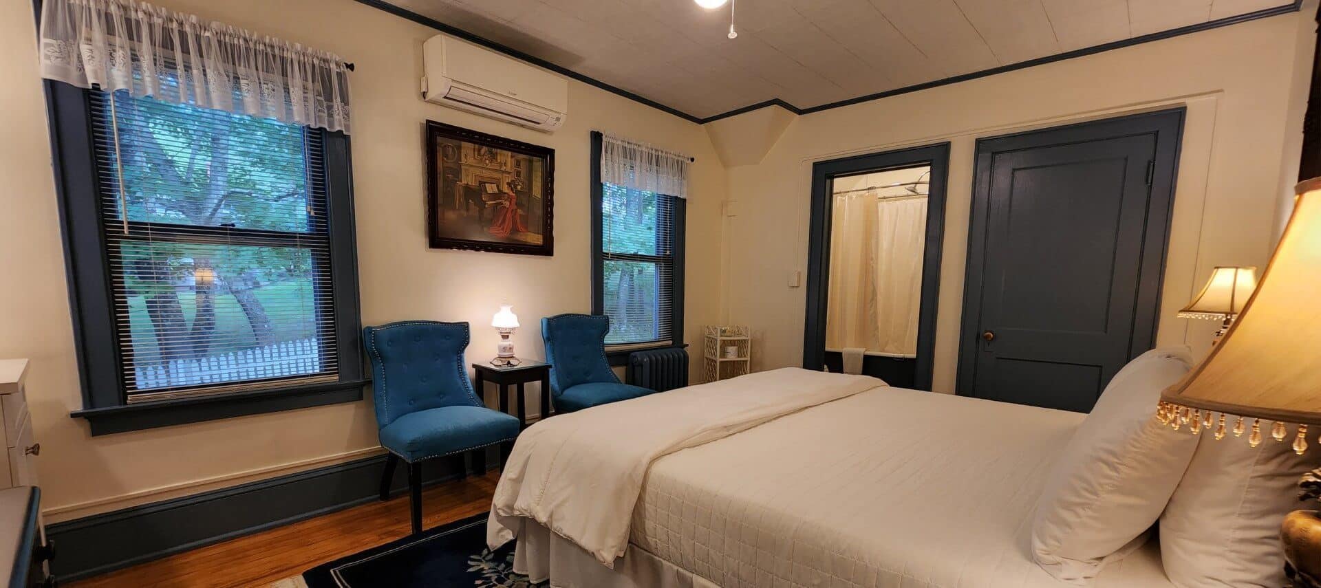 Guest room with bed in white linens, dark blue accents, two sitting chairs and large windows with lace valences
