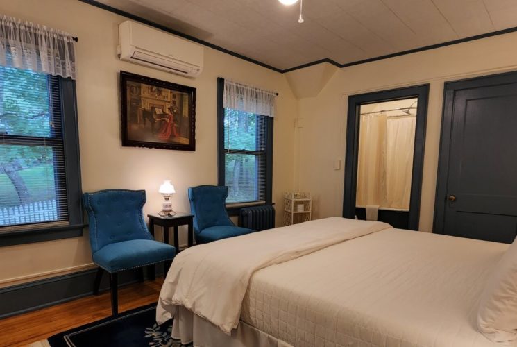 Guest room with bed in white linens, dark blue accents, two sitting chairs and large windows with lace valences