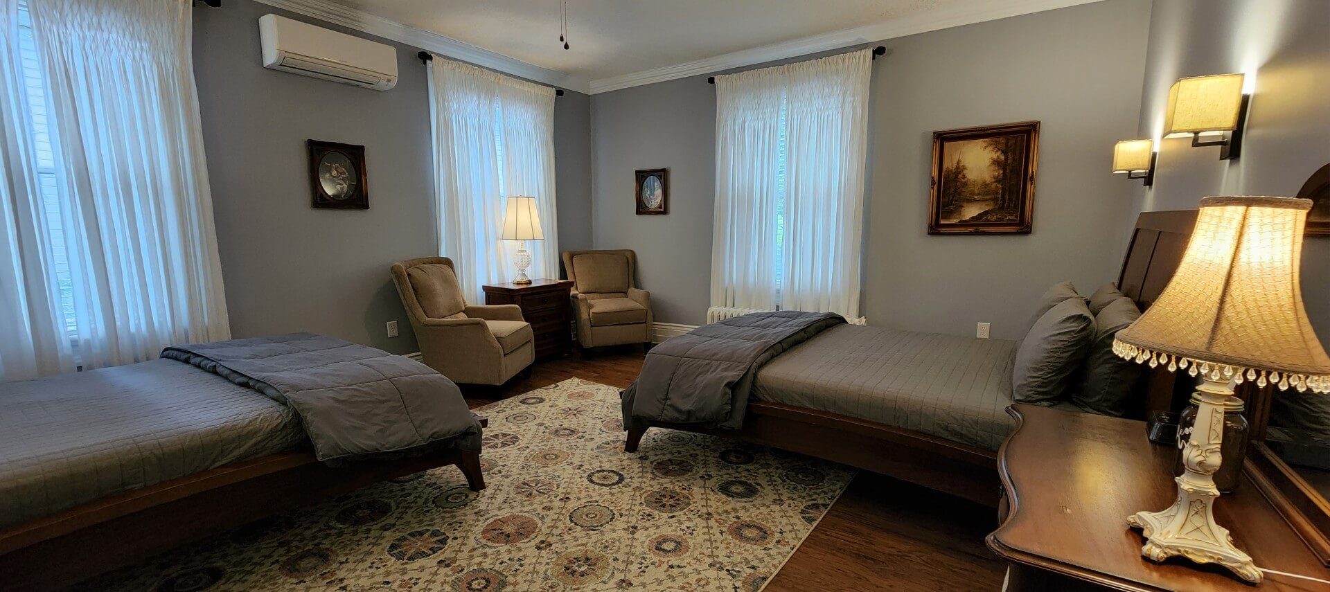 Guest room with two beds in grey linens, two sitting chairs and multiple soft-lit lamps