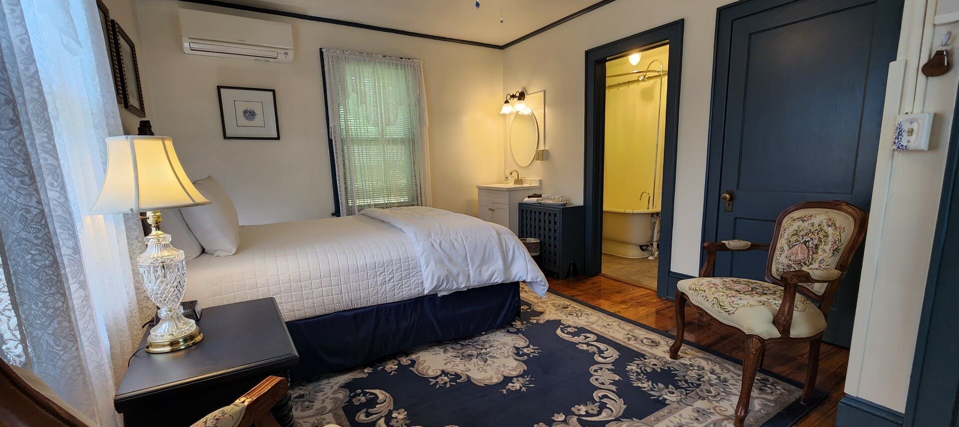 Guest room with bed in white linens, decorative blue rug and doorway into a bathroom with a clawfoot tub