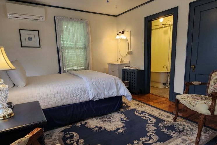 Guest room with bed in white linens, decorative blue rug and doorway into a bathroom with a clawfoot tub