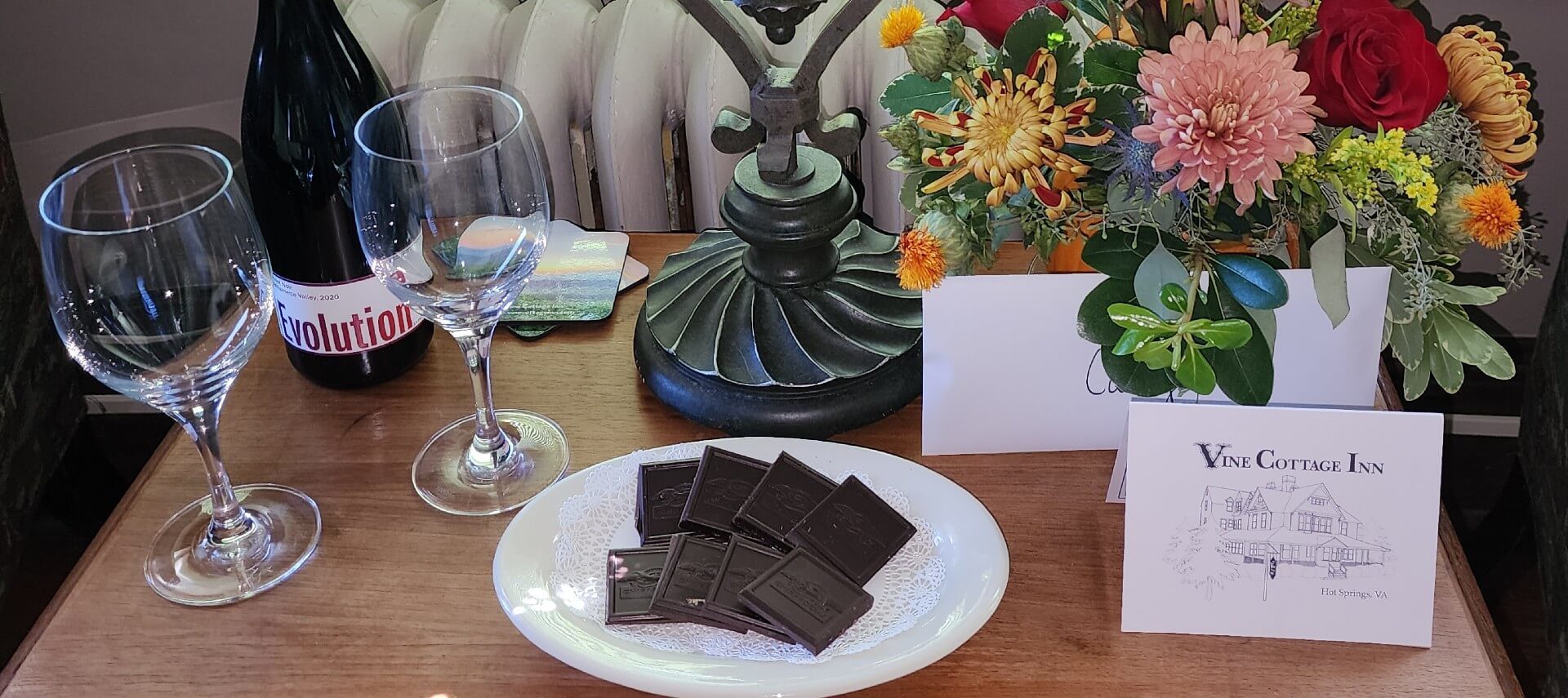 Table with wine and two glasses, vase of flowers with a card and plate of square chocolates