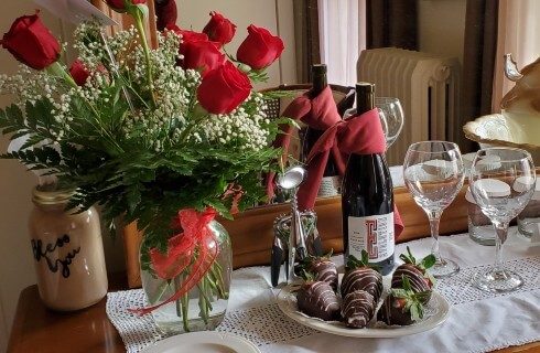 Table with vase of red roses, wine with two glasses and plate of chocolate covered strawberries
