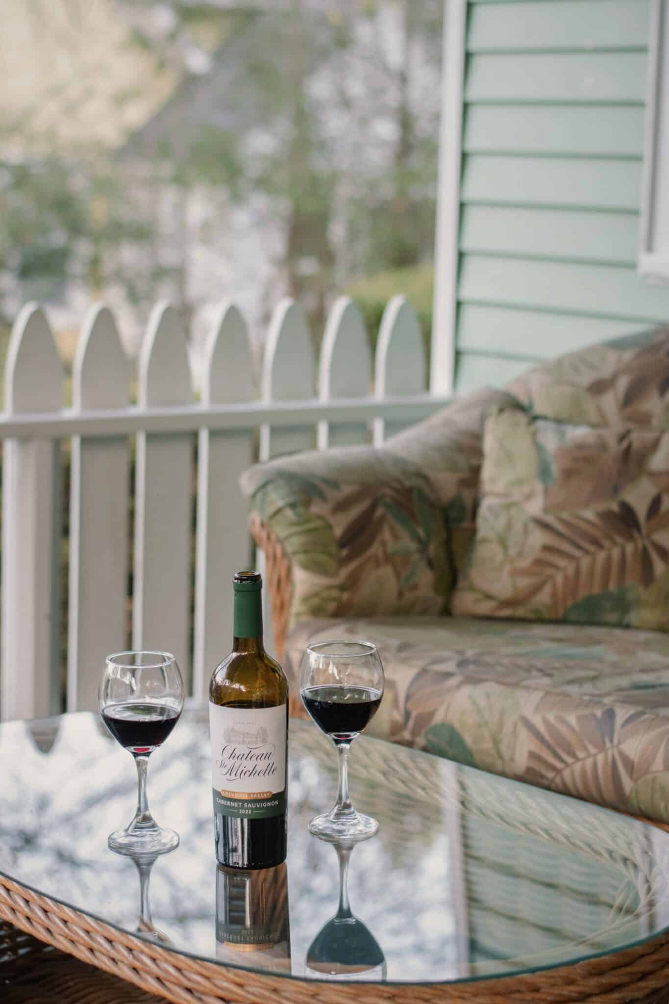 Bottle of red wine on a glass table with two wine glasses, outdoor porch setting at Vine Cottage Inn with a wicker chair nearby